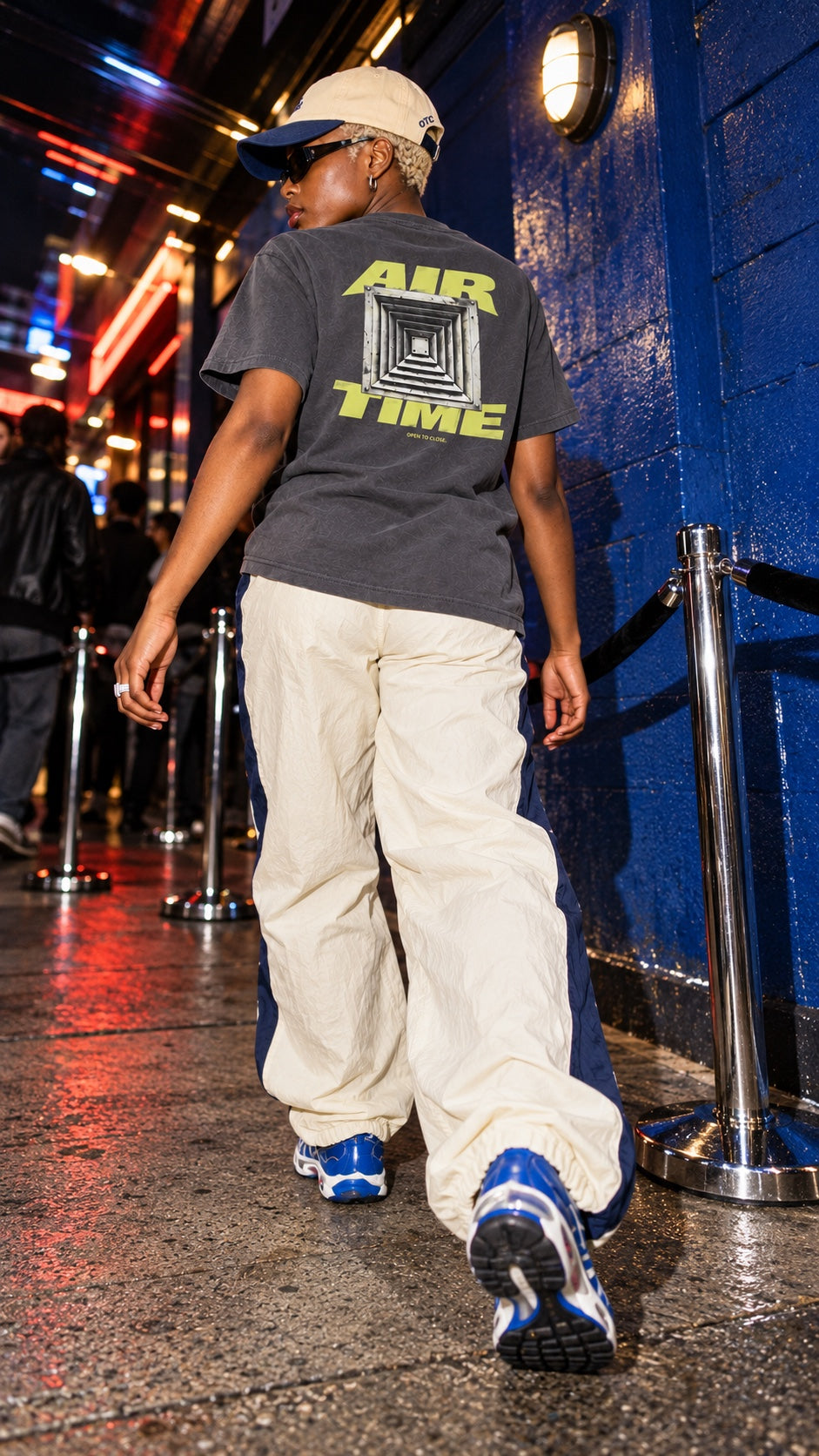 Air Time Tee worn by a model, back view, with the a/c tourist cap in Navy/Khaki, in a queue by a blue wall at night