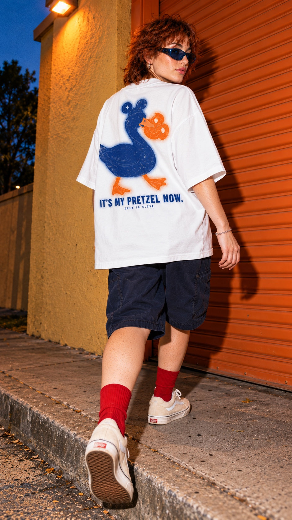 It’s My Pretzel Now Oversized Tee worn by a model, back view, by an orange shutter at dusk