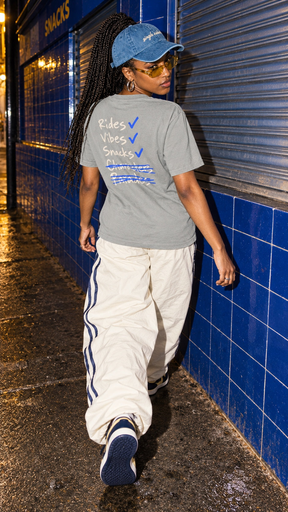 Rides, Vibes & Snacks tee worn by a model, back view, on a wet street by a blue-tiled wall at night