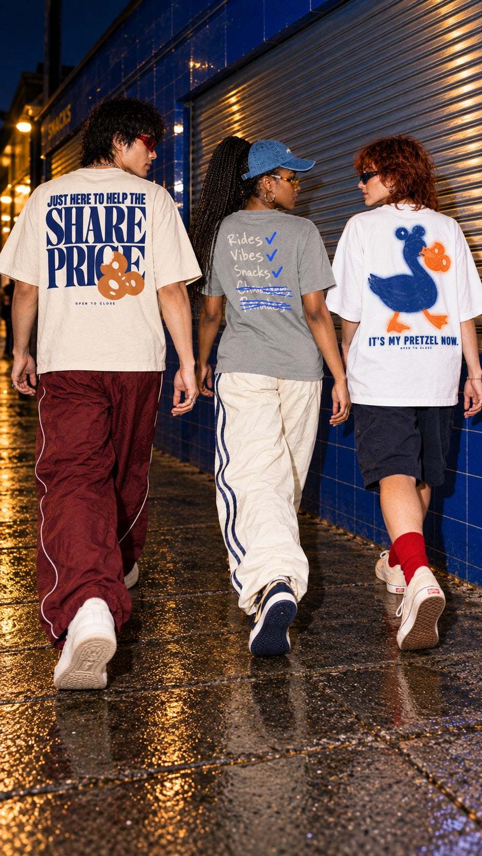 Three friends walking away on a wet street at night, back views: Share Price Boxy Tee (left), Rides, Vibes & Snacks Tee with Single Rider Denim Cap (centre), It's My Pretzel Now Oversized Tee (right)